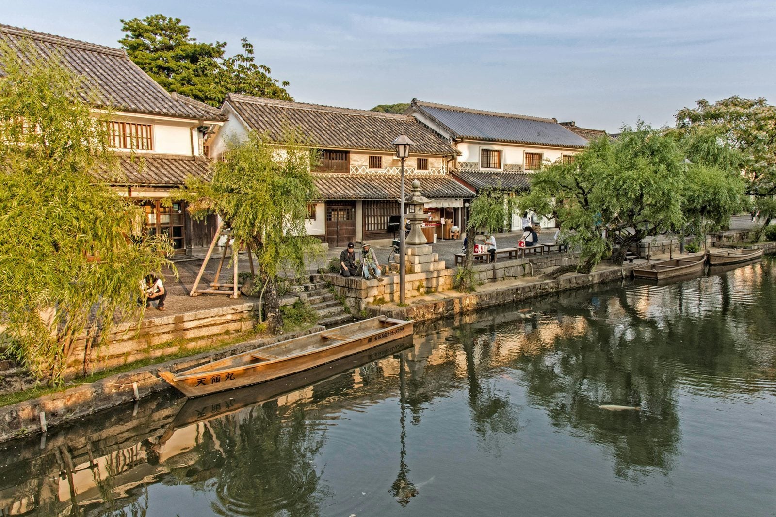 Stone-lined canal in the historic Kurashiki Bikan district, with willow trees, traditional white-walled storehouses and a wooden boat on the water